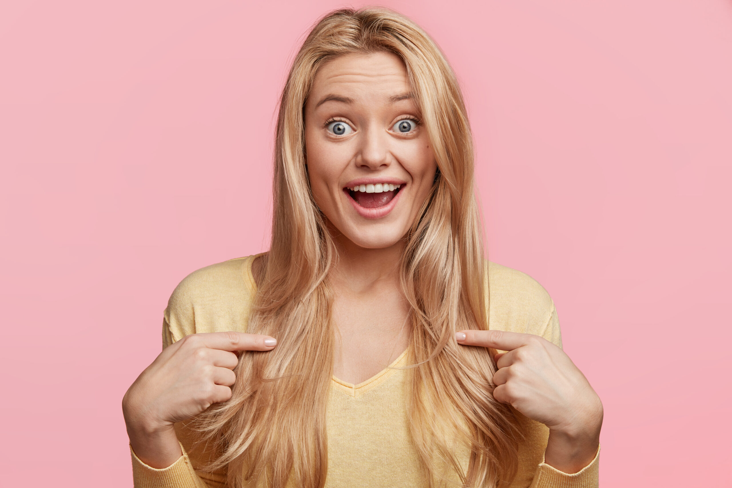 Lucky happy bugged eyed female student wears casual yellow sweater feels surprise, points at herself, can`t believe in good fortune, isolated over pink background. People and success concept optimistische junge Frau